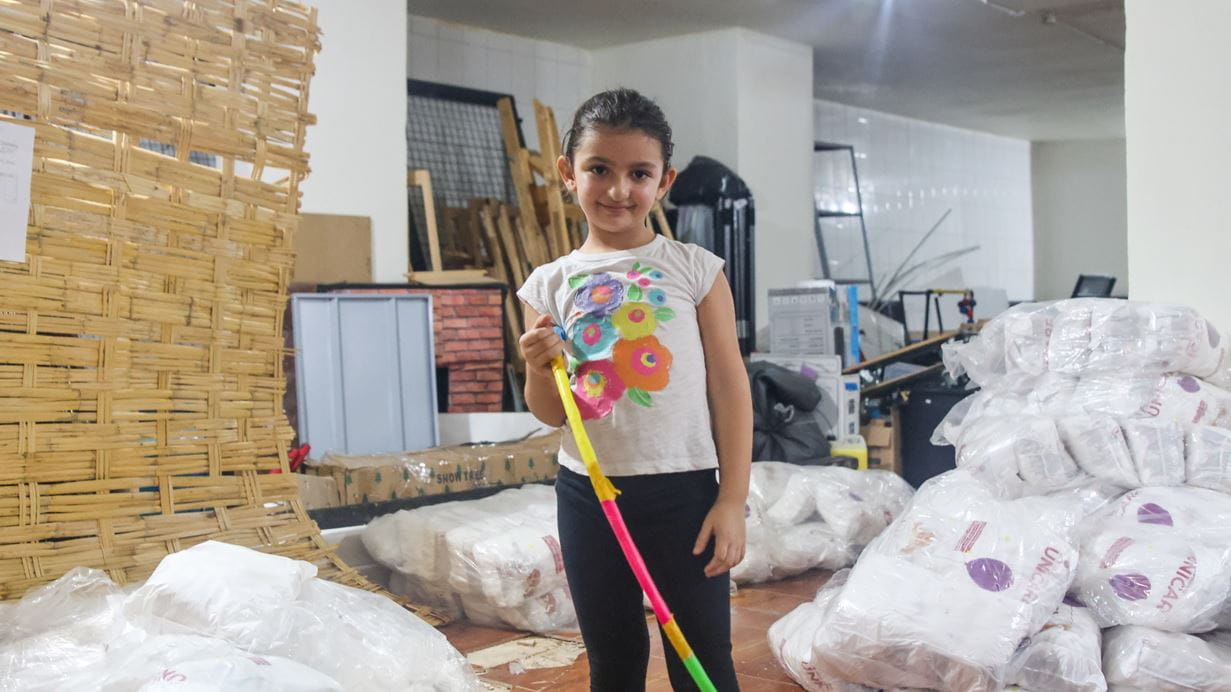 A young Lebanese girl with hair in a ponytail, wearing a white T-shirt with flowers and dark leggings, holds a brightly-coloured hula hoop as she stands amid supplies prepared for families like hers who have been displaced by the fighting in the south of Lebanon near the border with Israel.