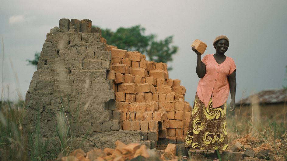 A woman is smiling as she stands beside a pile of bricks, holding a single brick