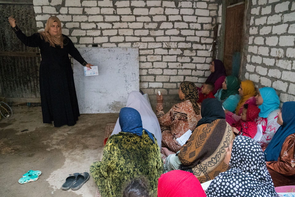 Teacher at board instructing literacy class of women seated on floor.