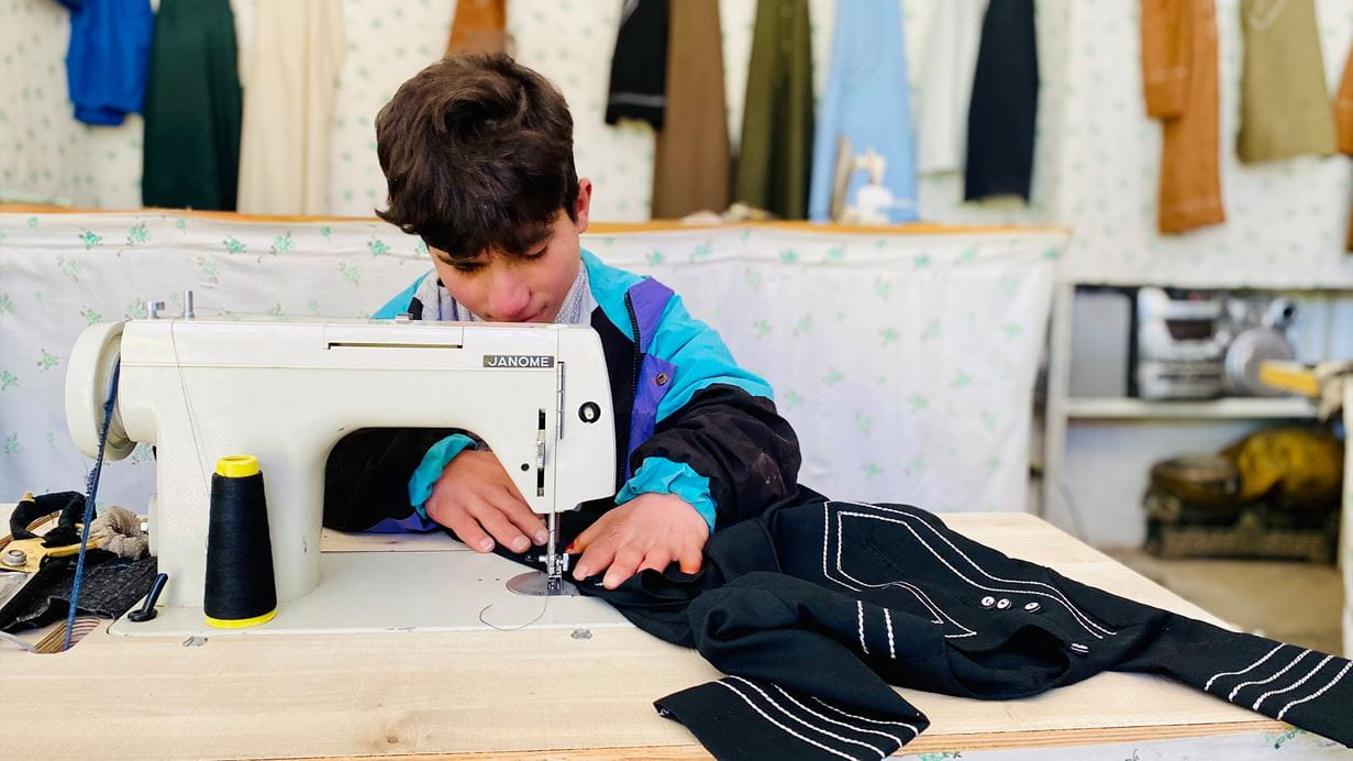 Young boy sewing black fabric on a Janome sewing machine, wearing a colorful jacket.