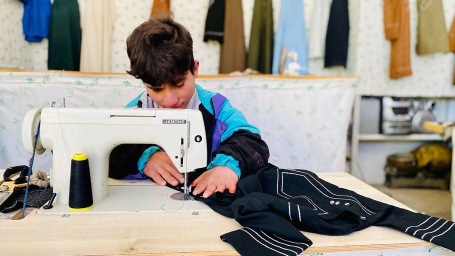 Young boy sewing black fabric on a Janome sewing machine, wearing a colorful jacket.