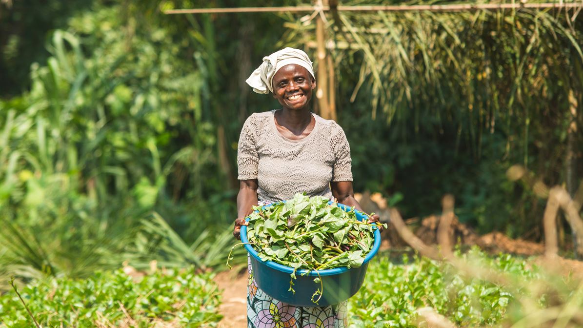 A smiling woman wearing a headscarf holds a large blue bowl of green vegetables in a lush outdoor garden.