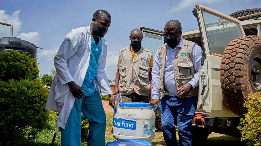 Three men carry a large white container labelled 'tearfund' with blue handles and lid. The man on the left wears a white lab coat, while the other two wear khaki utility vests. An off-road vehicle and a black water tank are visible in the background.