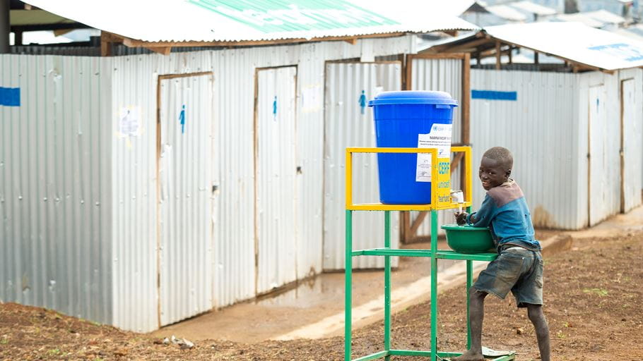 A boy washing his hands at a water tank, turns and smiles at the camera