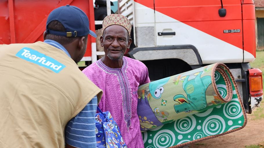 A Nigerian man in a purple tunic and patterned cap smiles while holding a rolled-up children's playmat, as another Nigerian man in a Tearfund vest looks on near a truck.