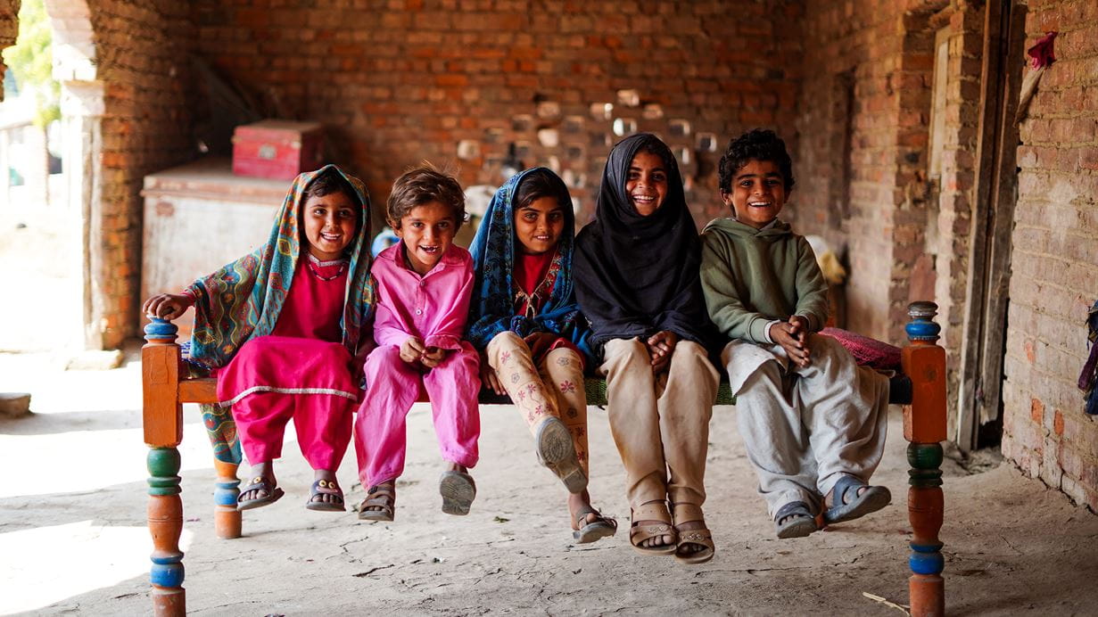 Five smiling children sit on a wooden bench in front of a brick wall.