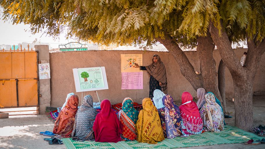 Women in colorful headscarves attend outdoor class led by woman pointing at chart under a tree.