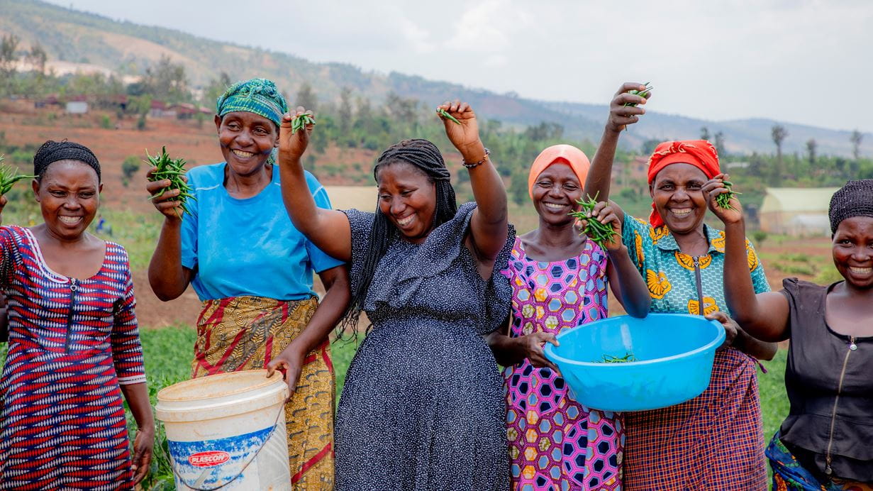 A group of Rwandan women stand outdoors, smiling and holding up small green vegetables in blue and white buckets. The background shows a hilly landscape.