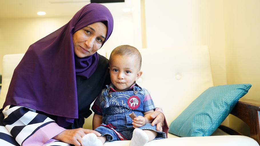 A woman dressed in a head covering puts her arm around her young child sat on a chair.