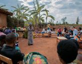A group of people sit on benches in a circle and woman stands in the middle, speaking to the group gesturing with her hands out at her side.