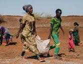 Two people in the foreground carry a heavy sack between them. Behind them is a barren landscape and other people.