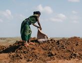 A woman is stood on a mound of soil on the ground and empties a sack of soil on top.