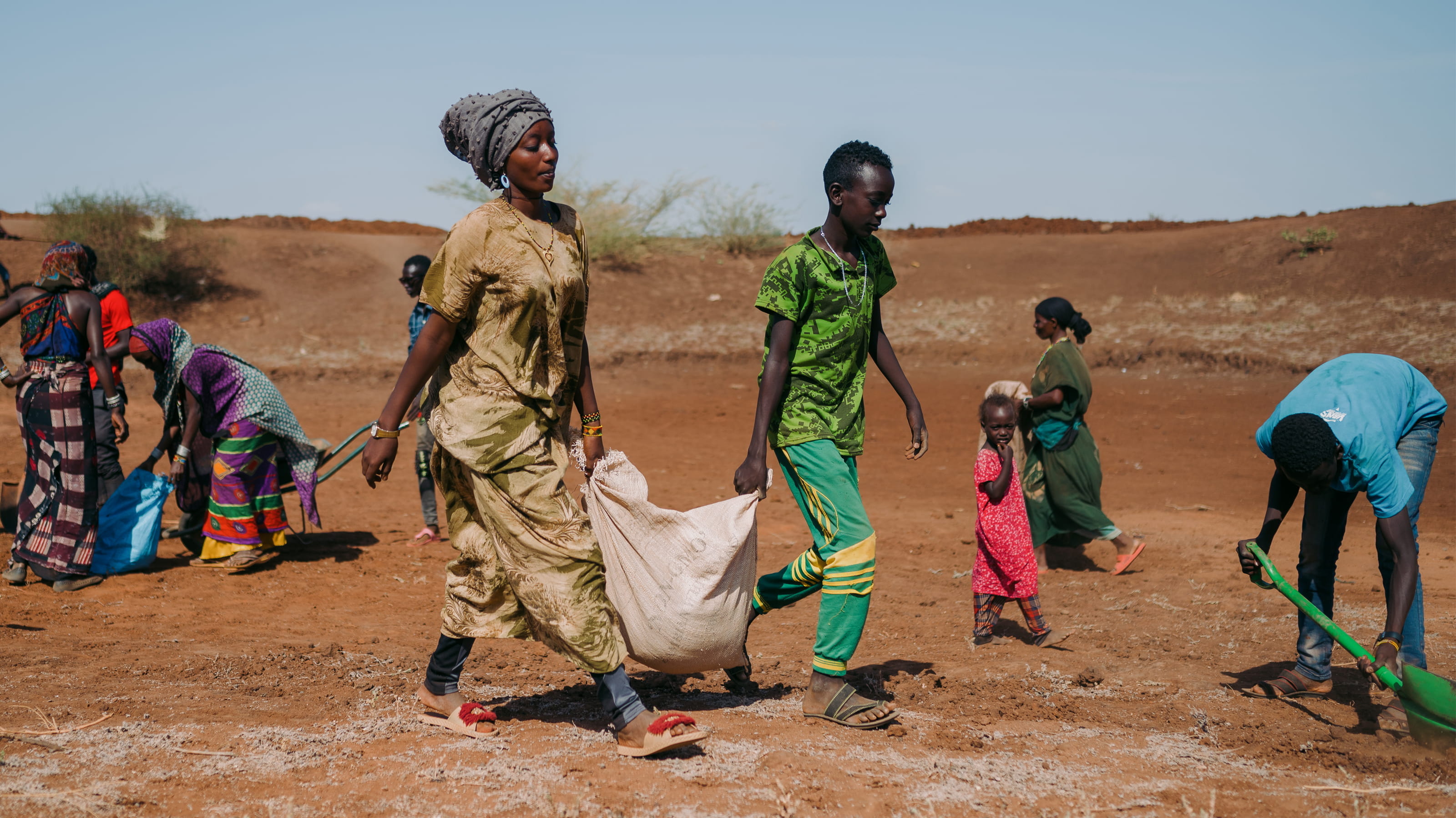 A woman and a boy carry a large sack between them. There are several people behind them, one is digging the ground.