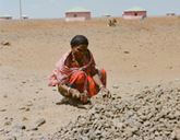 A woman is crouched on the ground with a tool in one hand and a rock in the other. In the foreground are lots of smaller stones.