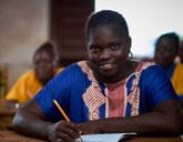 A woman sits at a desk with pen and paper and smiles toward the camera.