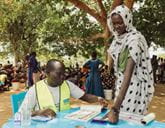 A male aid worker in a high-visibility vest sat at a desk with forms and a woman in a dotted headscarf gives her finger print, with people gathered under trees in the background.