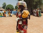 A woman carries a baby on her back and a large, colourful striped bundle on her head. She stands in a dusty outdoor area with other people and sacks visible in the background.