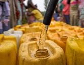 Close up of a hose filling a yellow water container, with lots of containers behind out of focus.