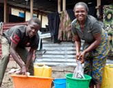 A woman and boy crouch over buckets of water as they wash clothes. They both smile at the camera.