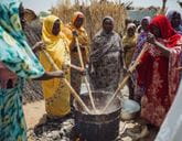 Group of women in colorful headscarves stirring a large pot of food cooking over an outdoor fire.