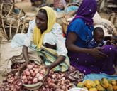 Woman in yellow headscarf selling onions in a bustling market, another woman with child sits nearby.