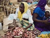 A woman crouches and holds a bowl of vegetables at a market.