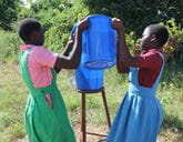 Two girls wearing pinafore dresses pour water from one large blue bucket to another.