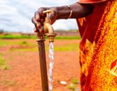 A metal tap in a dry barren field flows with water, a hand rests on top of the tap.