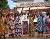 A group of eight women stand outdoors, holding hands above their heads, wearing brightly patterned dresses and skirts.