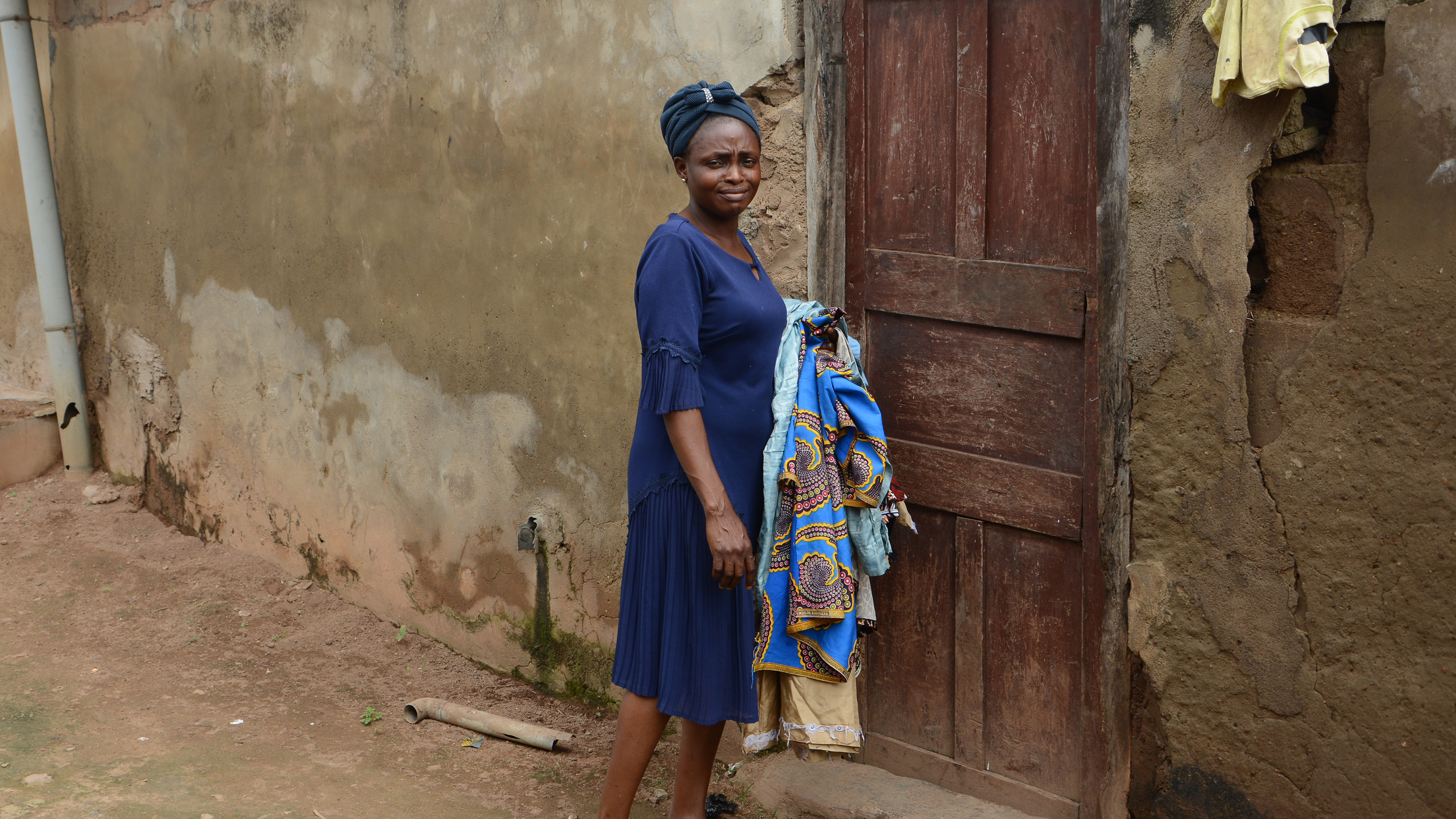 A woman stands outside near a wooden door and turns back to look at the camera. She holds fabric with her left arm.