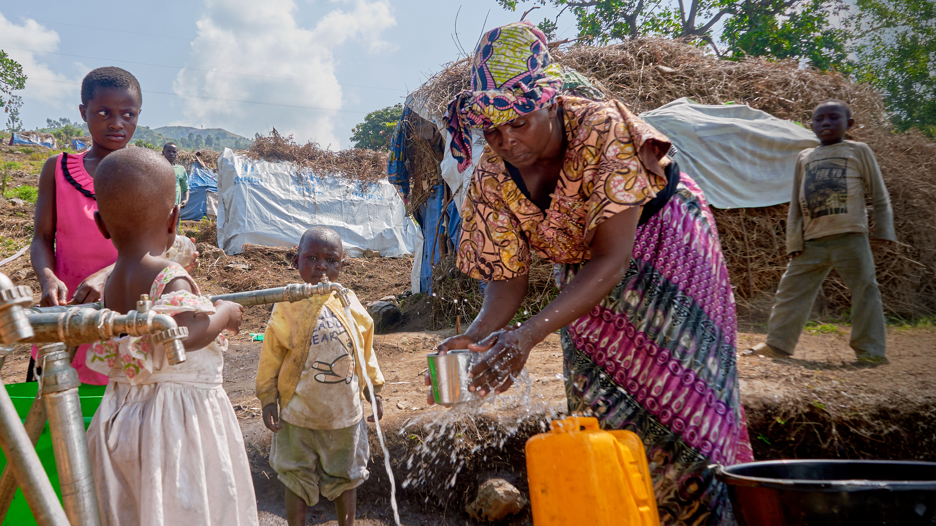 A woman bends over a water tap to fill a metal cup. Beside her is a large water bottle and several children.