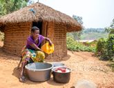 A woman pours water from a yellow container into a metal bowl, in another bowl there are several dishes. Behind her is a straw and mud hut.