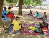 A group of people on blankets on the floor and two people on chairs sit under a tree in a circle.