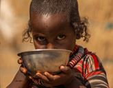 A child holds a small metal bowl up to their mouth and looks at the camera.