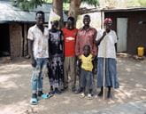 A family all stand together smiling in front of some trees and small buildings.