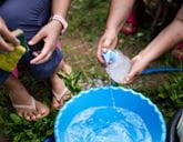 Two people crouch near a blue bucket of water, one person holds sponges and the other person fills the bucket with liquid soap.