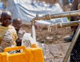 A yellow jerrycan is being filled with water from a tap, with children blurred in background.