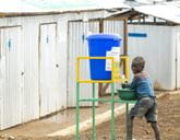 A young boy stands near a water tank with a tap to wash his hands, and looks back at the camera to smile.