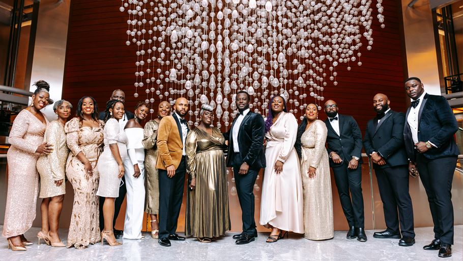 A large group of smiling Black men and women in formal gowns, suits, and tuxedos, posing in front of a wooden wall and a shimmering chandelier made of glass droplets.