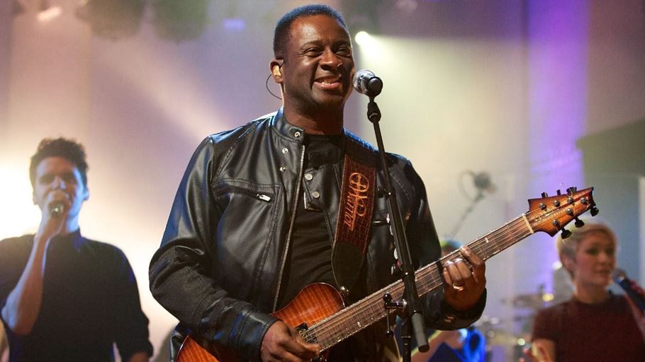 A man in a leather jacket singing into a microphone and playing an acoustic guitar on a stage. Behind him are two people singing into microphones.