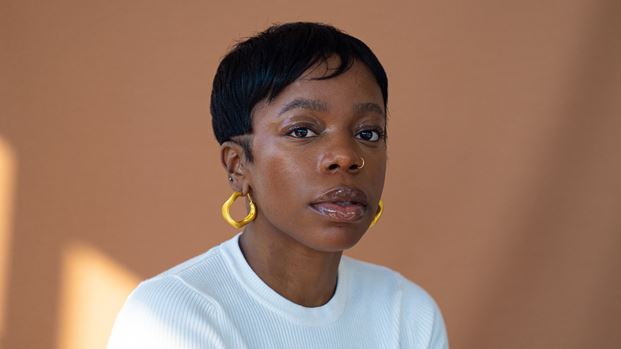 A headshot of a woman with short black hair with a shaved line above the ear, a gold nose ring, and large gold wavy hoop earrings. She is wearing a white ribbed top against a plain brown background.