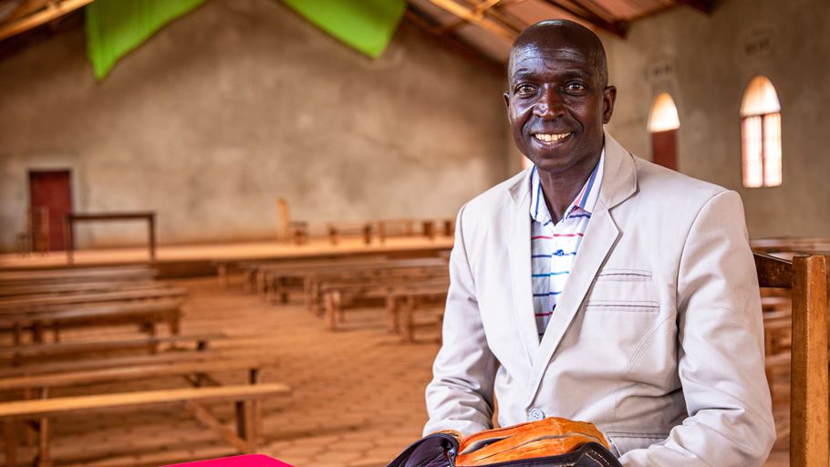 A man is sat in a church building, holding an open bible and smiling at the camera