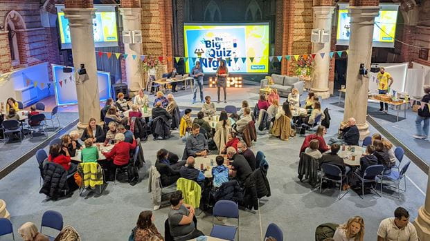 Photo of a church congregation in a spacious building participating in a quiz event. The room is decorated with colorful banners, and people are sitting at tables with pens and paper, looking engaged and excited. A podium with a big screen reading 'Big Quiz Night' is visible at the front of the room.