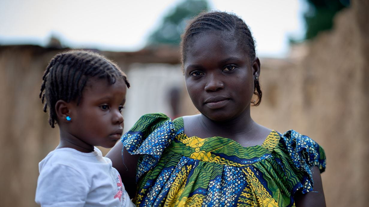 Davilla in colourful patterned clothing stands holding her daughter in her arms.