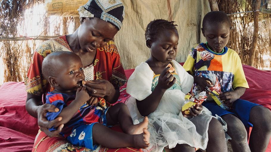A woman feeds a baby in her lap while two other young children sit beside them, eating. They are inside a simple shelter with woven walls.