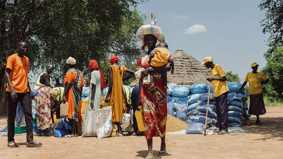 A woman carries a baby and a bundle on her head, walking past large stacks of blue sacks and a thatched hut, with other people gathered in a dusty outdoor area.