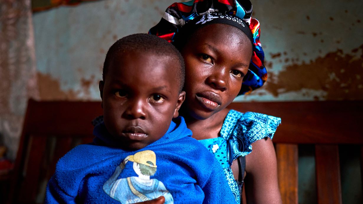 A young woman holds her child on her lap, both looking intently at the camera.