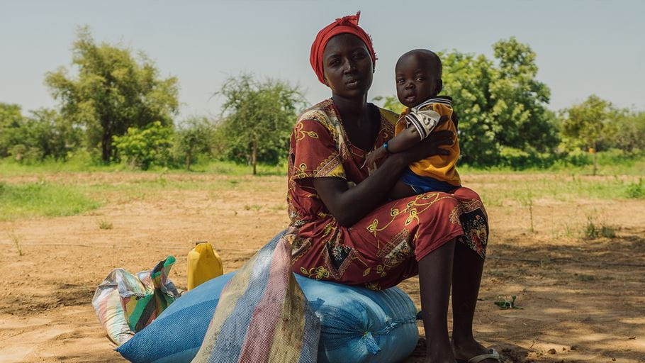 A woman in a red patterned dress and headscarf sits on blue sacks, holding a baby in a yellow and blue outfit. They are outdoors on dry earth with sparse trees and green foliage in the background. A yellow jerry can and another striped sack are next to them.