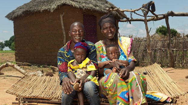 A man and woman are smiling and holding two small children outside a hut.