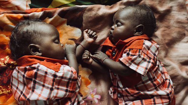 Two babies lie side-by-side on a patterned blanket, wearing matching orange and white checked shirts.